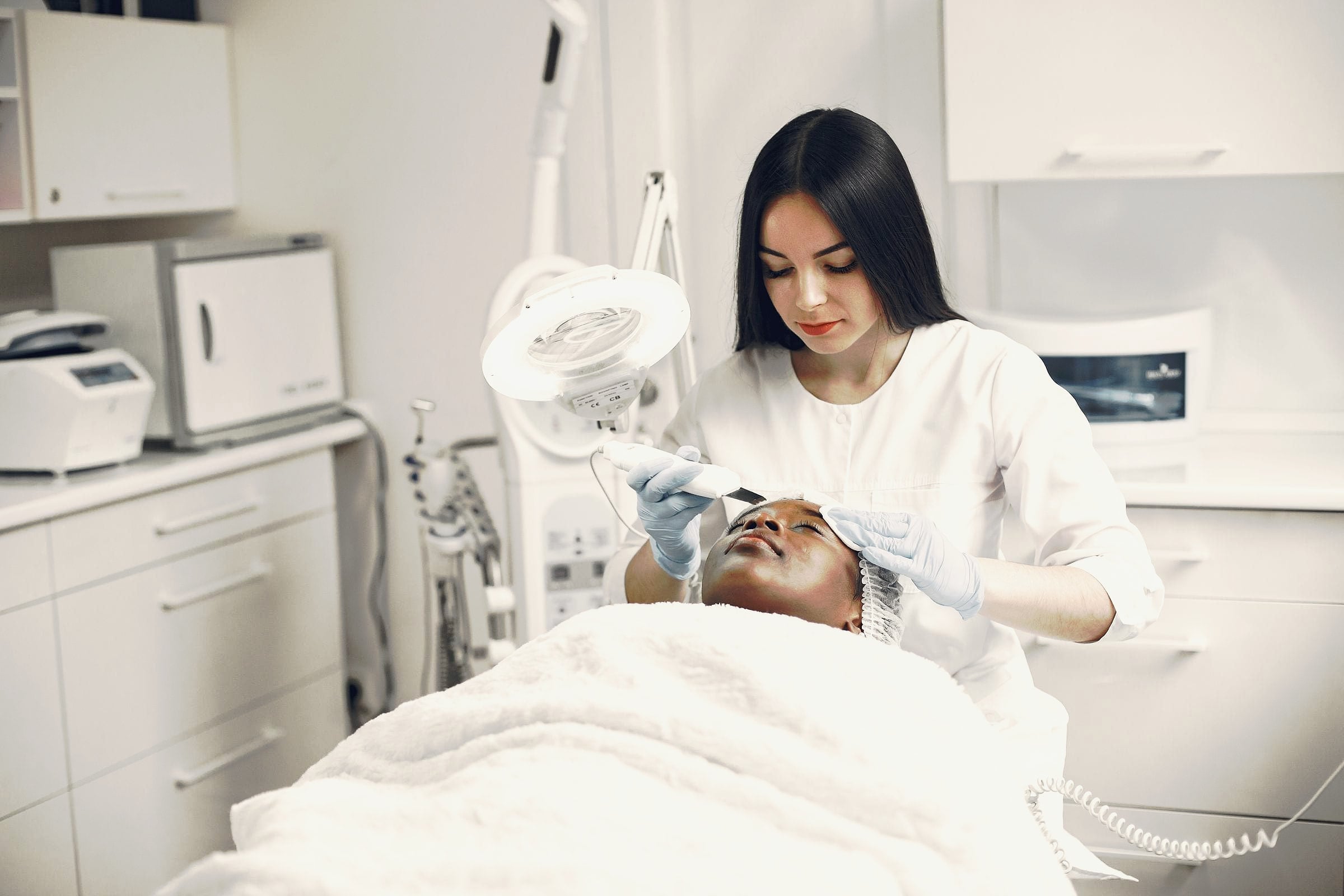 An aesthetician giving a client a facial treatment with a handheld device in a bright treatment room.