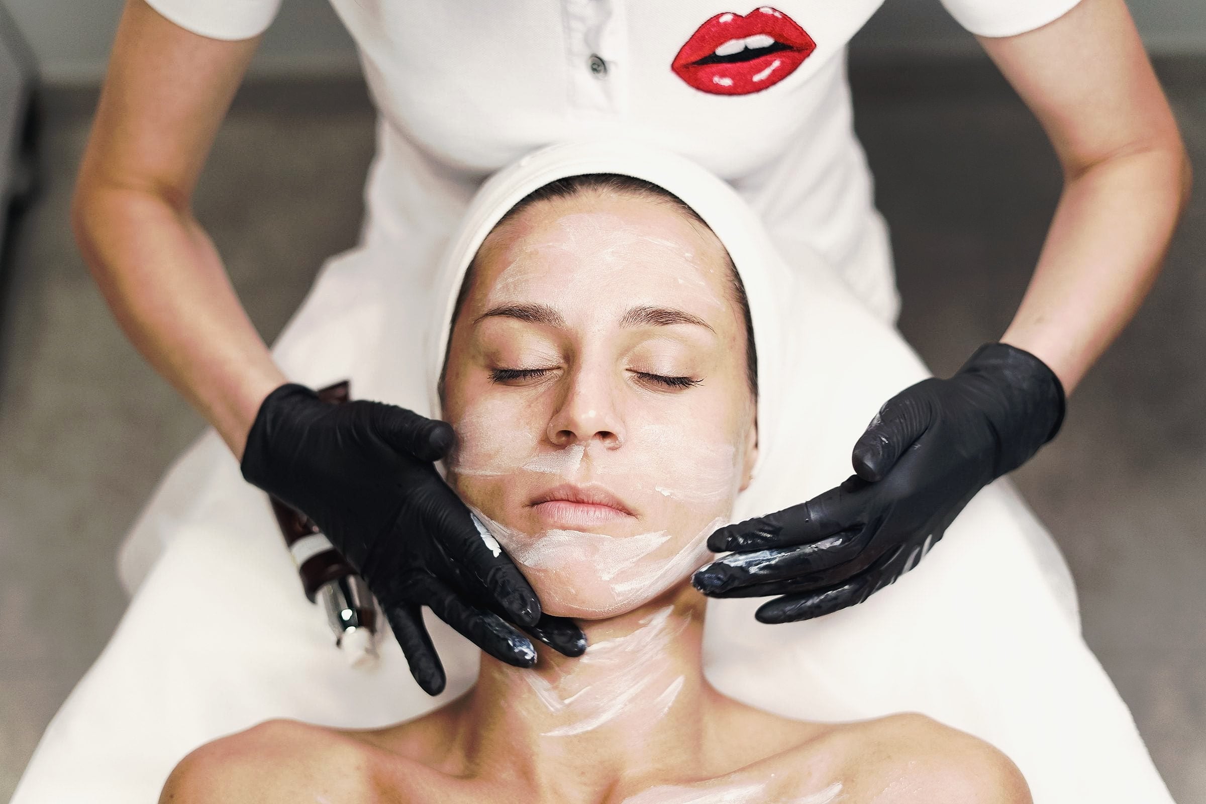 A beautician in black gloves applying a facial mask to a relaxed client.