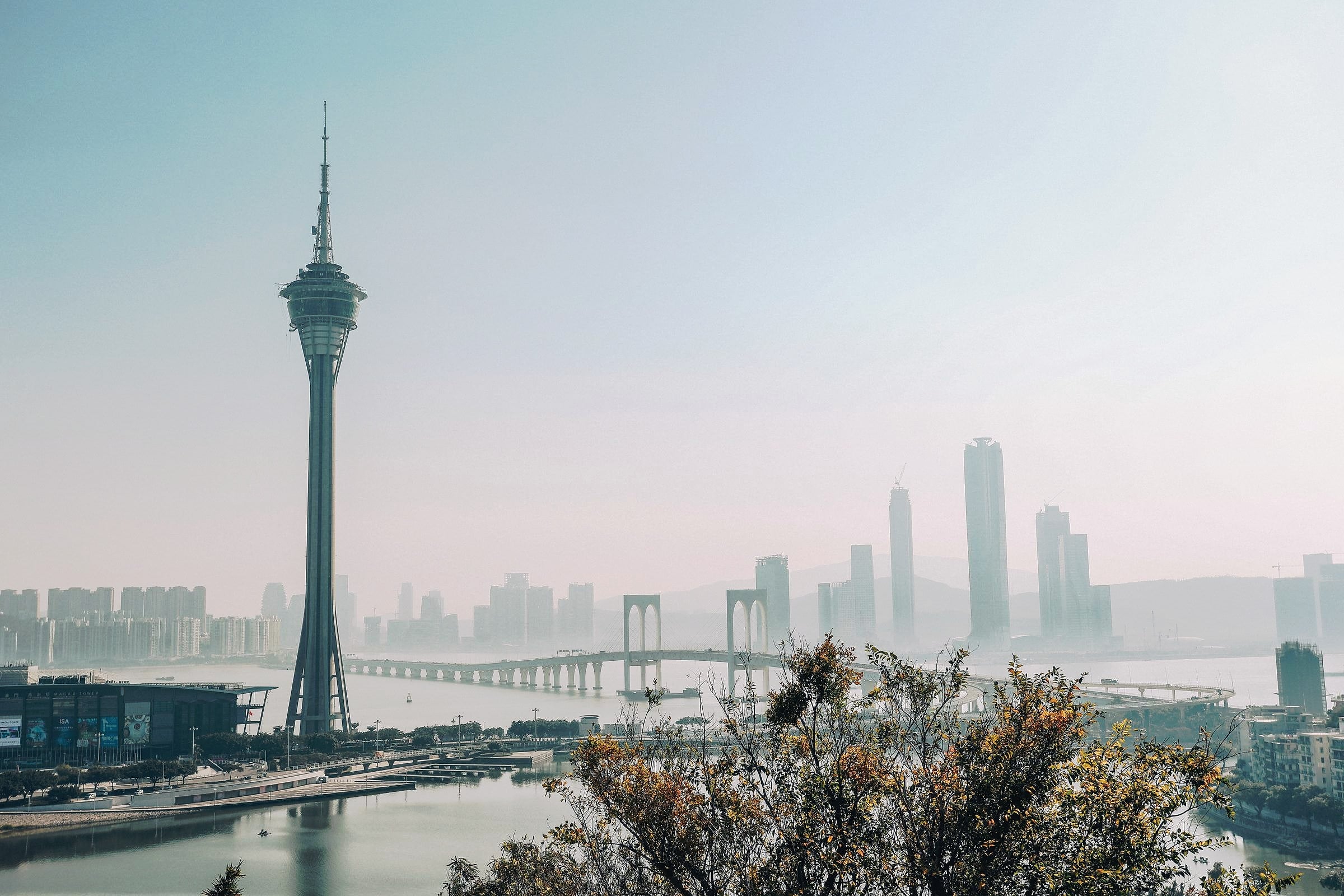 Macau Tower and the waterfront skyline under a clear sky, with a bridge across the water.