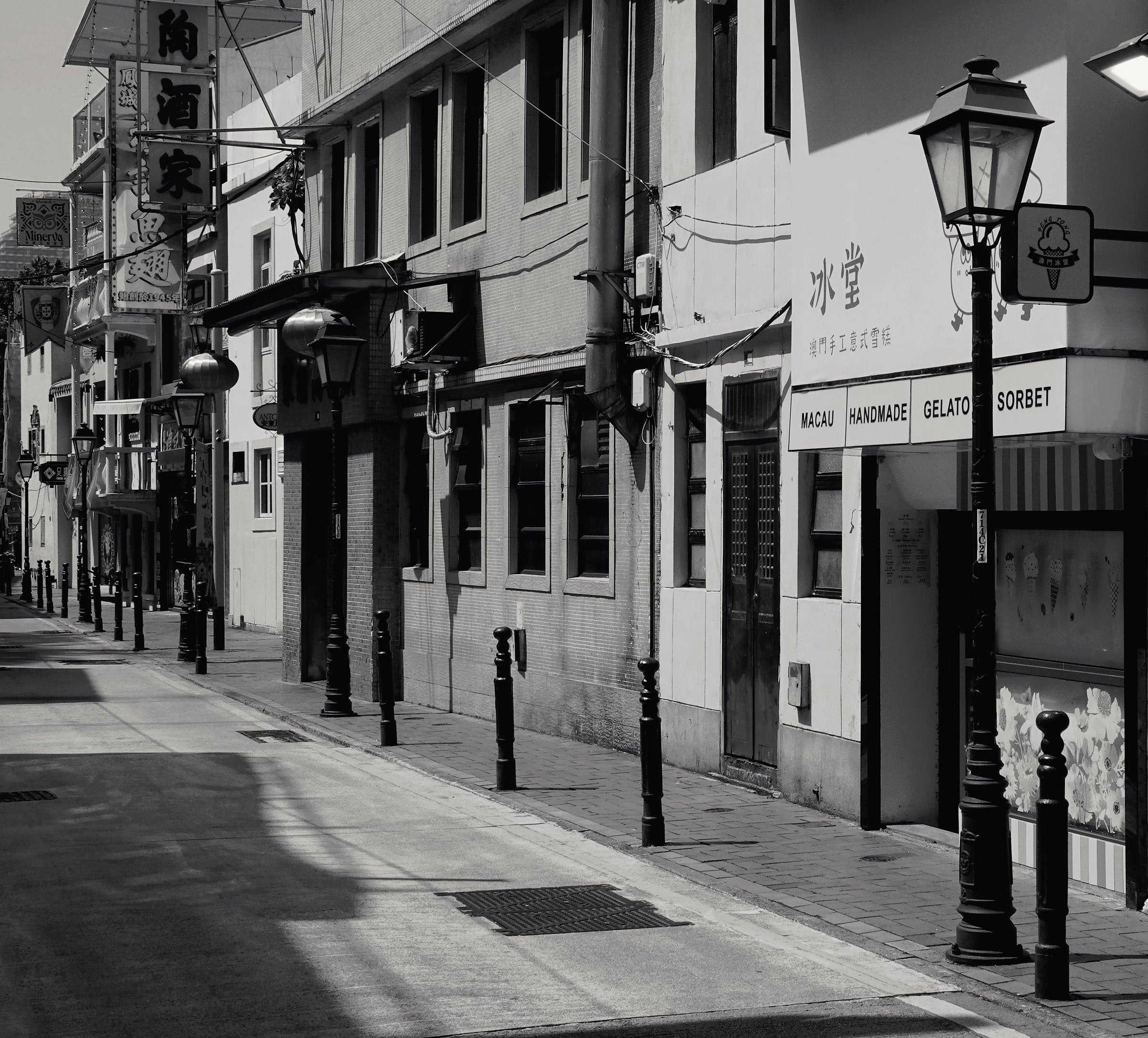 A quiet lane in Taipa Village with old shopfronts and iron street lamps, in black and white.